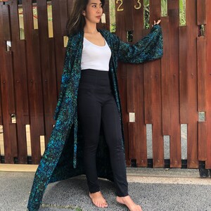 May include: A woman wearing a long, teal and black patterned kimono with a white tank top and black pants. She is standing in front of a wooden fence with the number "23" on it.
