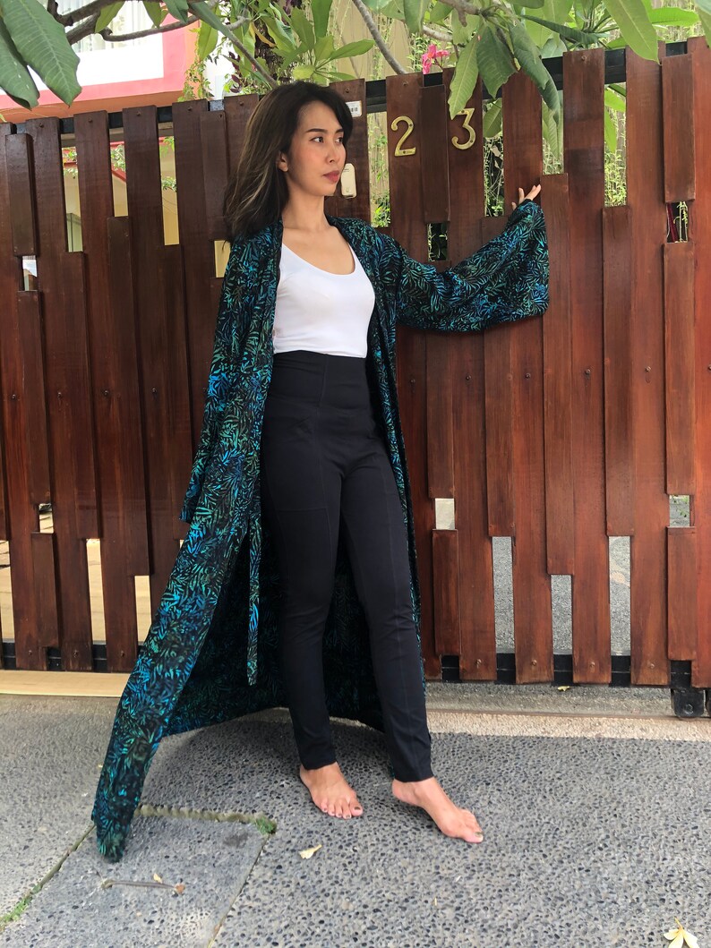 May include: A woman wearing a long, teal and black patterned kimono with a white tank top and black pants. She is standing in front of a wooden fence with the number "23" on it.