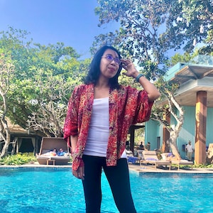 May include: A woman wearing a red and yellow patterned kimono with a white tank top and black pants. She is standing by a swimming pool with a building in the background.