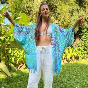 May include: A blue and white patterned kimono with a white background and blue and purple polka dots. The kimono has a fringe trim and is worn over white pants.