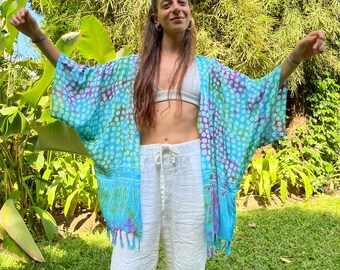 Pode incluir: Um quimono estampado em azul e branco com fundo branco e bolinhas azuis e roxas. O quimono tem uma borda de franja e é usado sobre calças brancas.
