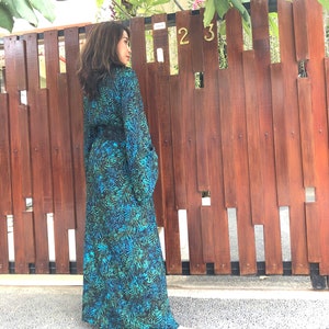 May include: A woman wearing a long, blue and green patterned dress with a black belt. The dress has a long sleeve and a V-neck. She is standing in front of a wooden fence with the numbers "23" on it.