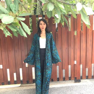 May include: A long, black and green patterned robe with a white top underneath. The robe has long sleeves and a loose fit. The person is standing in front of a wooden fence.