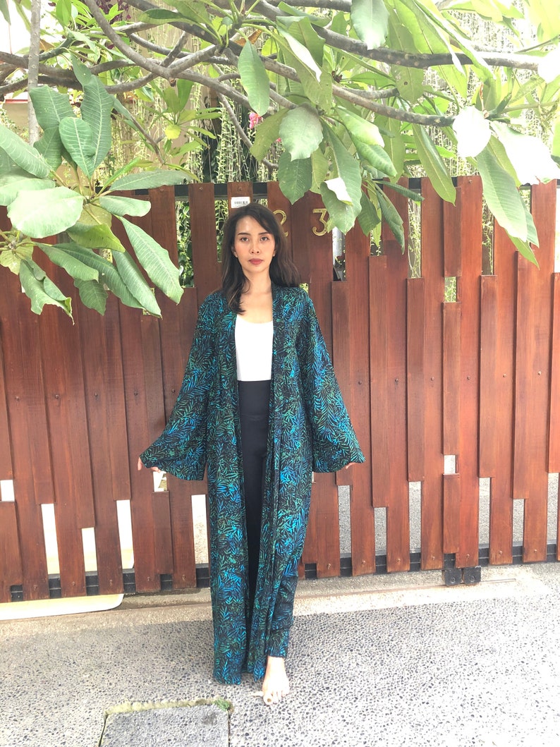 May include: A long, black and green patterned robe with a white top underneath. The robe has long sleeves and a loose fit. The person is standing in front of a wooden fence.