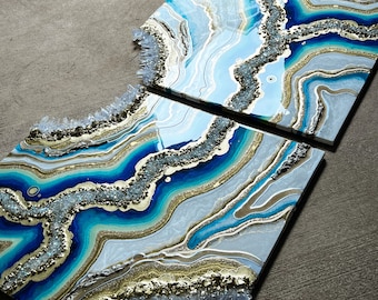 May include: Two abstract resin art pieces with a blue, white, and gold agate design. The pieces are set against a gray background. The top piece is an arch shape and the bottom piece is rectangular.