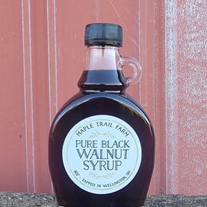 May include: A clear glass bottle filled with dark syrup, featuring a black cap and a small handle. The label reads "Maple Trail Farm Pure Black Walnut Syrup" with the text "8oz - Tapped in Wellington, OH."