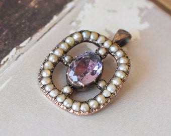 May include: An antique pendant featuring an oval, light purple gemstone at the center, surrounded by small, round pearls. The pendant has a decorative, openwork design with a bronze-colored metal frame. The piece is likely vintage or antique.