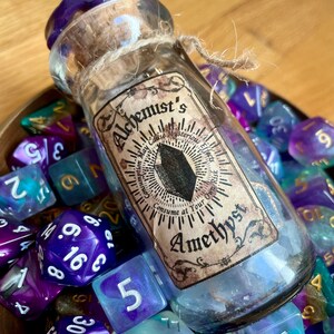 Peut inclure: Une petite bouteille en verre avec un bouchon violet et un bouchon en liège. La bouteille est remplie d'un liquide clair et porte une étiquette sur laquelle est écrit "Alchemist's May Cause Mysterious Effects Consume at Your Own Risk Amethyst". La bouteille est entourée de dés irisés violets et bleus.