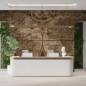 May include: A white reception desk with a gold trim sits in front of a wall with a vintage world map wallpaper. The map is in shades of brown and beige and has a puzzle-like design. Green plants are visible on either side of the desk.