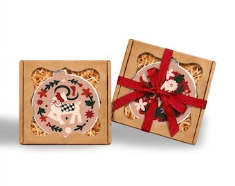 May include: Two square brown cardboard gift boxes with scalloped window cutouts, each containing a round ornament nestled in wood shavings. One ornament features a stylized deer, while the other is tied with a red ribbon and floral pattern.