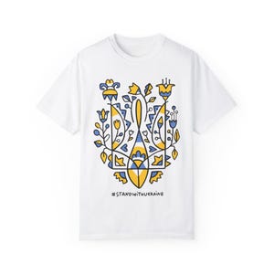Puede incluir: Camiseta blanca con un diseño floral estilizado azul y amarillo del tridente ucraniano. El texto '#STANDWITHUKRAINE' está impreso debajo del diseño.