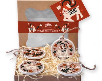 May include: A brown cardboard gift box containing three round wooden ornaments with folk-style illustrations of a deer, a bird, and floral patterns. The box features a red band with the text "gifty" and "Christmas decor" and a matching card.