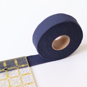 May include: A roll of dark blue bias binding, a type of fabric trim, is partially unrolled and lying on a white surface next to a ruler.