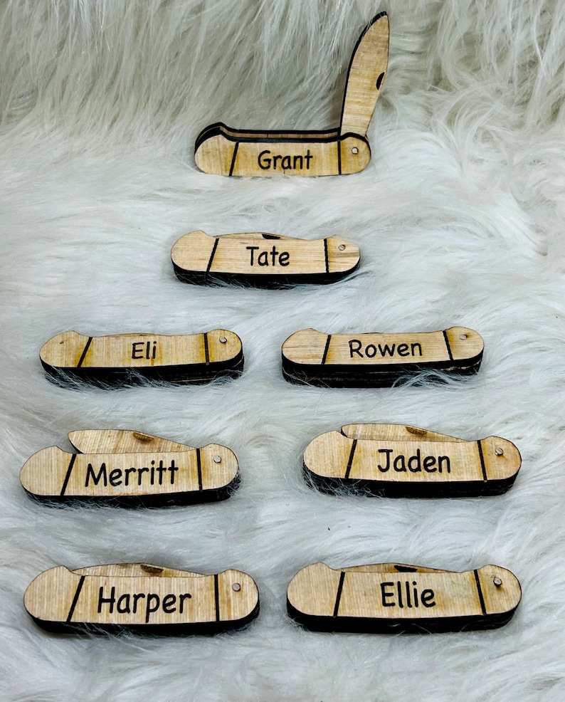 May include: Wooden pocket knife-shaped name tags with black accents. Each tag features a different name: Grant, Tate, Eli, Rowen, Merritt, Jaden, Harper, and Ellie. The tags are arranged on a fluffy white surface.