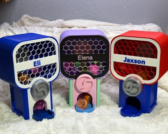 May include: Three colorful gumball machines, each with a unique design. The machines are blue, purple, and red, with the names "Eli", "Elena", and "Jaxson" displayed. Each machine has a honeycomb-patterned window.