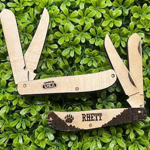 May include: A wooden pocket knife with a bear paw design and the name "Rhett" engraved on the handle. The knife is made in the USA.