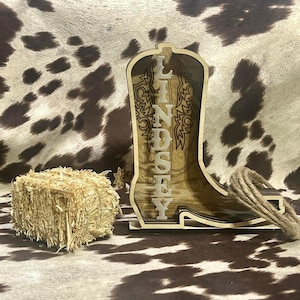 May include: Wooden cowboy boot cutout with the name Lindsey carved into the side. The boot is sitting on a brown and white cowhide background.