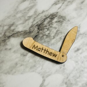 May include: A wooden pocket knife with the name "Matthew" engraved on the handle. The knife is light brown and has a blade that is partially open, revealing the knife's design. The background is a marble surface.