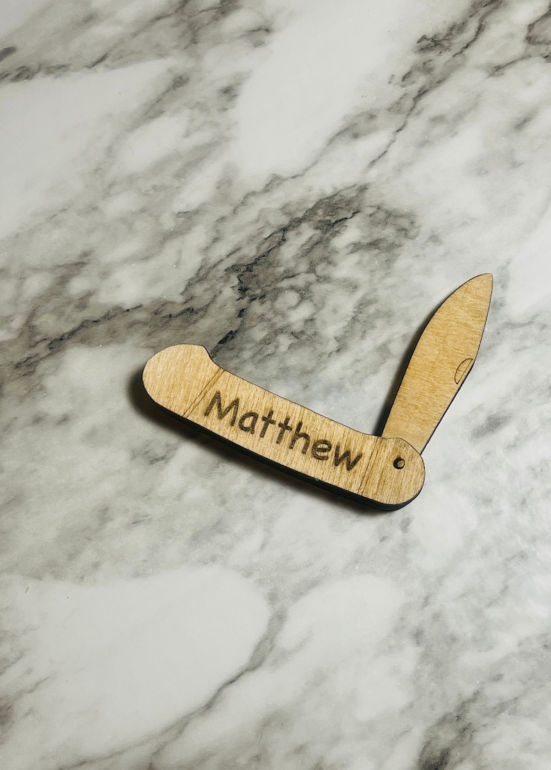 May include: A wooden pocket knife with the name "Matthew" engraved on the handle. The knife is light brown and has a blade that is partially open, revealing the knife's design. The background is a marble surface.