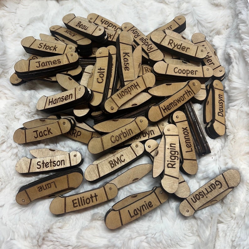May include: A collection of wooden pocket knife-shaped name tags. Each tag is engraved with a different name in a sans-serif font. The tags are made of light-colored wood and have a natural finish. The names include: Stock, James, Knox, Beau, Ryder, and others.