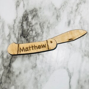 May include: A wooden folding knife with the name "Matthew" engraved on the handle. The knife is light brown and has a blade that is partially open. The background is a marble pattern.