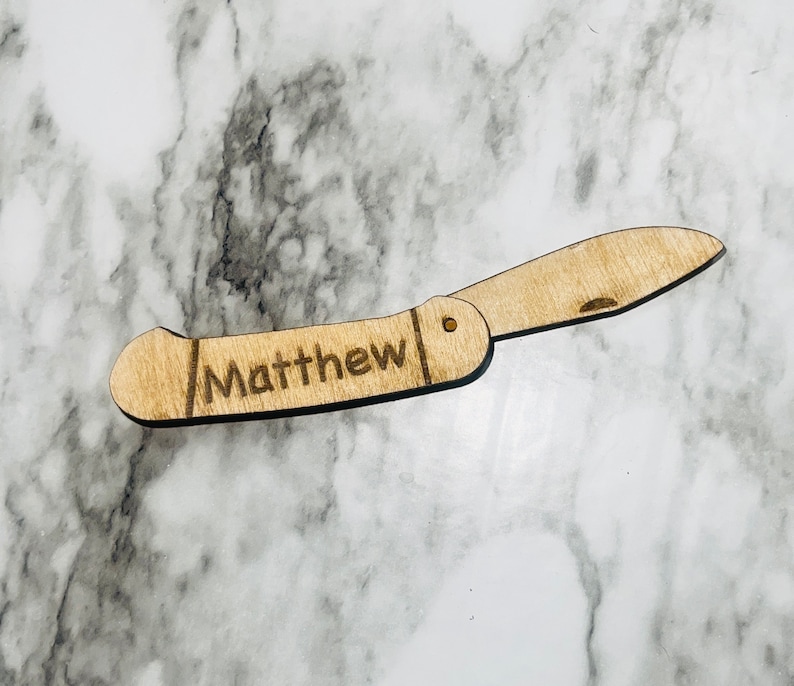 May include: A wooden folding knife with the name "Matthew" engraved on the handle. The knife is light brown and has a blade that is partially open. The background is a marble pattern.