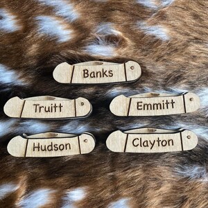 May include: Five wooden pocket knife-shaped name tags. Each tag is engraved with a different name: Banks, Truitt, Emmitt, Hudson, and Clayton. The tags are light brown and have a small hole on one end.