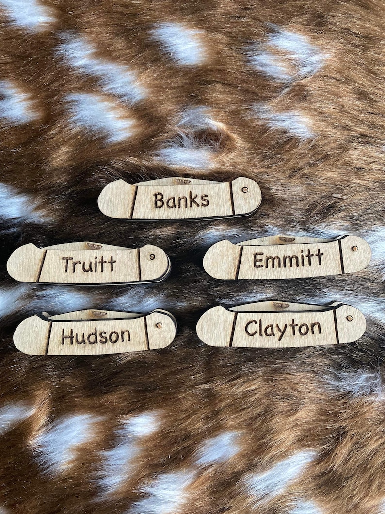 May include: Five wooden pocket knife-shaped name tags. Each tag is engraved with a different name: Banks, Truitt, Emmitt, Hudson, and Clayton. The tags are light brown and have a small hole on one end.