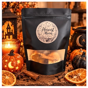 May include: A black stand-up pouch with a clear window at the bottom, featuring a circular label that reads Harvest Moon Simmer, decorated with illustrations of a crescent moon, orange slices, and cinnamon sticks.