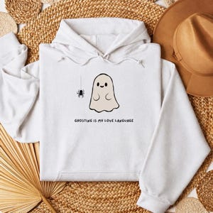 May include: A white hooded sweatshirt featuring a graphic of a small beige ghost and a black spider hanging from a thread. Below the graphic, the text reads GHOSTING IS MY LOVE LANGUAGE in black capital letters. The hoodie has long sleeves.