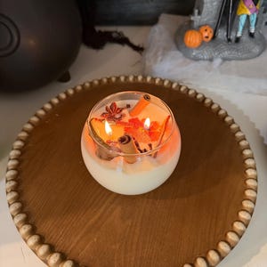 May include: A round glass candle with two lit wicks, filled with white wax and topped with decorative dried orange slices, cinnamon sticks, and star anise, resting on a circular wooden tray with a beaded edge.