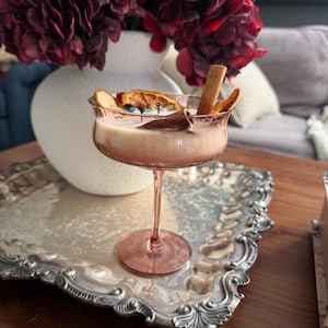 May include: A candle poured into a pink-tinted stemmed glass coupe. The wax is off-white and topped with dried orange slices and two cinnamon sticks. The glass has a wide, shallow bowl and a slender stem, resting on a circular base.