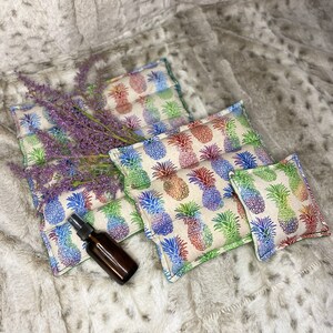 May include: Three rectangular pillows with a pineapple print in shades of green, blue, and purple. The pillows are arranged in a pyramid shape on a white faux fur surface. A small brown glass bottle is in front of the pillows.