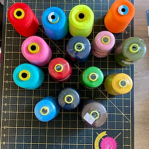 May include: A collection of 16 spools of thread in various colors, including red, blue, green, yellow, pink, and brown. The spools are arranged on a black cutting mat with a grid pattern. The spools are all the same size and shape, and they are all wrapped in thread.