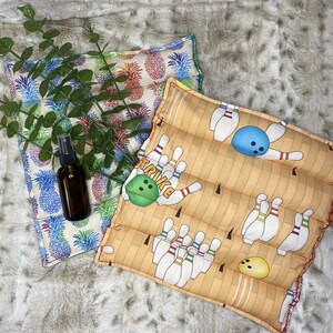 May include: A tan and brown fabric bowling-themed hot pack with a white and blue bowling ball, a yellow bowling pin, and the word "Strike" in black. The hot pack is on a white and gray fuzzy surface with a green plant and a clear glass bottle with a brown liquid.