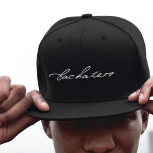 May include: A black, flat-brimmed baseball cap with a white embroidered cursive script that reads "bachatero".