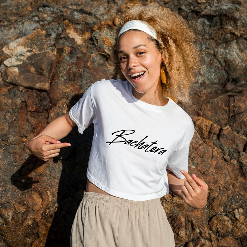 May include: A woman wearing a white crop top with the word "Bachatera" written in black cursive. She is smiling and pointing at the shirt. She has curly blonde hair and is wearing a white headband.