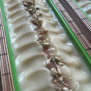 Może przedstawiać: Zbliżenie na ręcznie robioną kostkę mydła o kremowobiałym kolorze i pofałdowanej powierzchni. Suszone kwiaty jaśminu są rozrzucone po wierzchu mydła. Mydło znajduje się w zielonej prostokątnej formie z drewnianymi bokami.