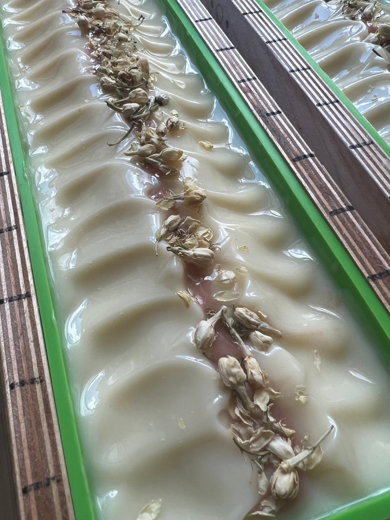 Może przedstawiać: Zbliżenie na ręcznie robioną kostkę mydła o kremowobiałym kolorze i pofałdowanej powierzchni. Suszone kwiaty jaśminu są rozrzucone po wierzchu mydła. Mydło znajduje się w zielonej prostokątnej formie z drewnianymi bokami.