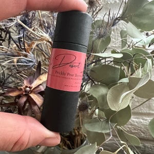 May include: A black tube with a pink label that reads "Desert Prickly Pear Beeswax Balm" with ingredients listed below. The tube is held in a hand against a background of dried flowers.