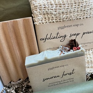 May include: A gift box set containing a green bar of soap labeled "gigglestars soap co. sonoran forest moisturizing & detoxifying Small Batch Handmade Soap", a natural loofah, and a wooden soap dish.