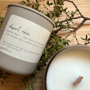 May include: Two candles with a wooden wick and the text "desert rain" on a label. The candles are gray and silver, surrounded by green sprigs on a wooden surface. The label includes the text "breathe deep...the dance of the desert".