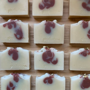 Może przedstawiać: Ręcznie robione kostki mydła z kremowo-białą bazą i bordowymi wirami. Każda kostka jest ostemplowana napisem "giggletars" i ułożona na drewnianej powierzchni. Niekt&oacute;re kostki mają złote listki. Kolekcja mydeł rzemieślniczych.