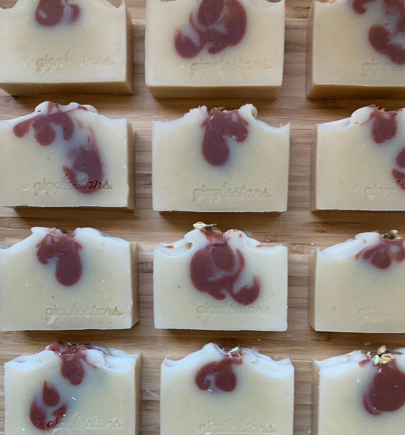 Może przedstawiać: Ręcznie robione kostki mydła z kremowo-białą bazą i bordowymi wirami. Każda kostka jest ostemplowana napisem "giggletars" i ułożona na drewnianej powierzchni. Niekt&oacute;re kostki mają złote listki. Kolekcja mydeł rzemieślniczych.