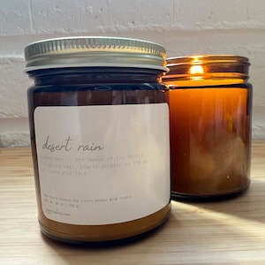 May include: A brown glass candle jar with a silver lid. The candle is labeled "desert rain" and has a description of the scent. The candle is sitting on a wooden surface.