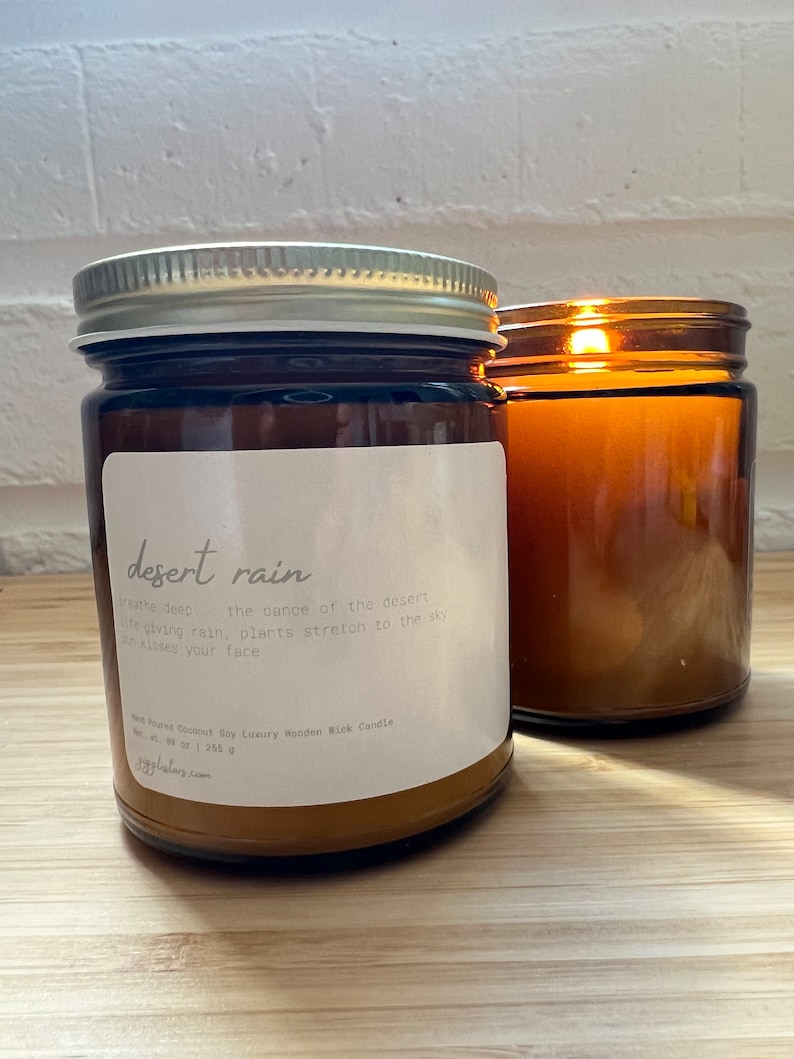 May include: A brown glass candle jar with a silver lid. The candle is labeled "desert rain" and has a description of the scent. The candle is sitting on a wooden surface.