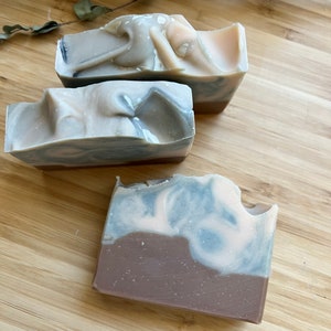 May include: Three handmade soap bars with a marbled design. The soaps feature layers of brown, light blue, and white, with a swirled pattern. The bars are rectangular and have a natural, organic aesthetic.
