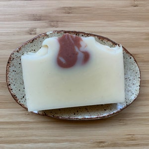 Może przedstawiać: Prostokątna kostka mydła w jasnokremowym kolorze z brązowym wirkiem, spoczywająca na nakrapianej ceramicznej mydelniczce. Mydelniczka ma brązową obw&oacute;dkę i leży na drewnianej powierzchni.