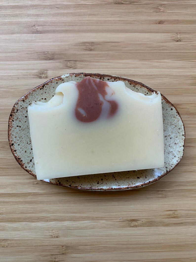 Może przedstawiać: Prostokątna kostka mydła w jasnokremowym kolorze z brązowym wirkiem, spoczywająca na nakrapianej ceramicznej mydelniczce. Mydelniczka ma brązową obw&oacute;dkę i leży na drewnianej powierzchni.