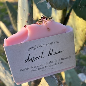 May include: A pink handmade soap bar with dried flowers and herbs on top. The label reads "gigglestars soap co. desert bloom Prickly Pear Cactus & Himalayn Rhubarb Small Batch Handmade Soap 5 oz | 142 g"